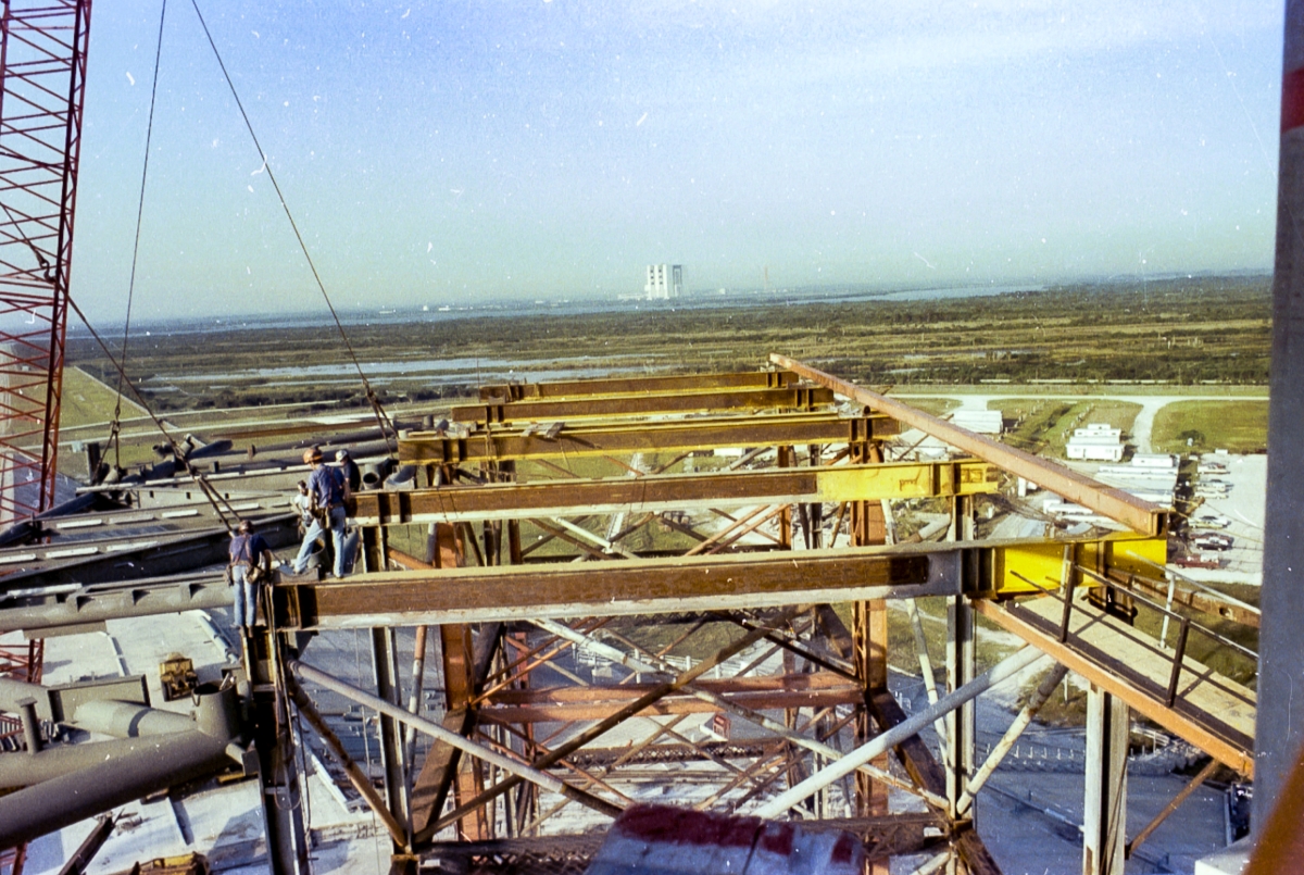 Large-format photograph: Wilhoit Steel Erectors has lifted the RSS Bottom Truss Weldment almost the full eighty some-odd vertical feet it needs to travel, to permit it to then be moved out and across the top of the Falsework which you see clearly in this image, taken from the FSS at Elevation 140. Note the Union Ironworkers from Local 808, none of whom are tied-off with any sort of safety gear of any kind. This is how ironworking was practiced in 1980 by the best in the business, and they all despised each and every attempt by safety operatives to ensnare them in any kind of ropes, lines, harnesses, or anything else which could restrict their completely free motion on high steel. You simply walked out on the iron, went where you needed to go, and did your job once you got there. Photo courtesy Eugene Hajdaj.